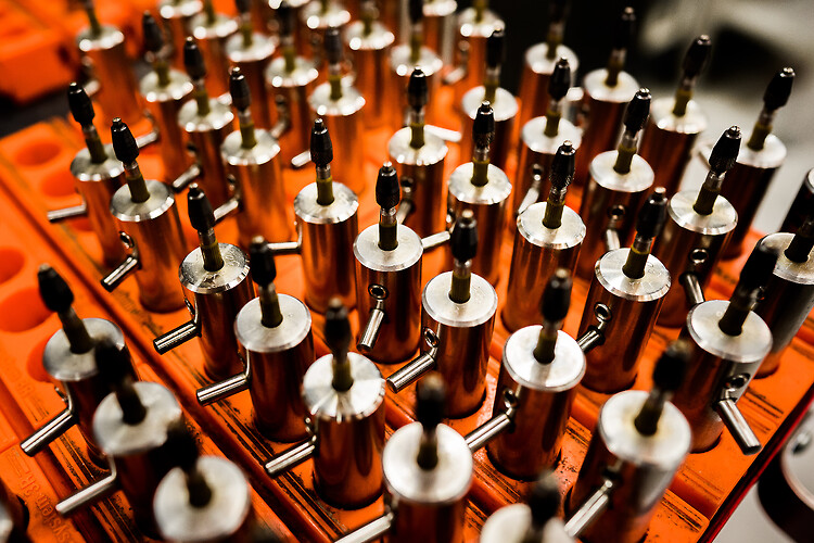 A high-angle, close-up shot of rows of cylindrical metal electrode holders arranged in a grid within an orange industrial tray. The silver holders each feature a horizontal locking pin and secure a vertical, dark-tipped tool, with a shallow depth of field keeping the central rows in sharp focus while blurring the foreground and background.